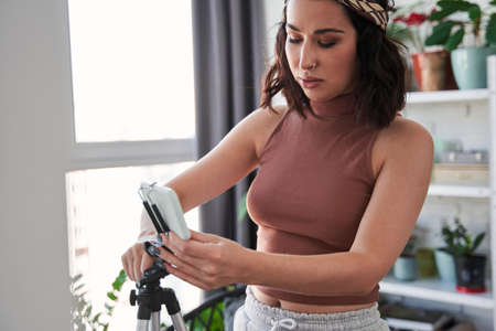Concentrated female blogger putting smartphone at the tripod while preparing shooting her blog
