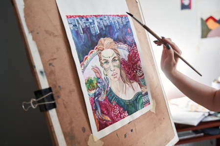 Female artist standing next to the easel and holding brush and artist palette while drawing her...