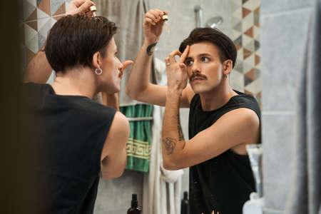 Calm metrosexual man is looking into mirror and putting some treatment from bottle for skin beauty