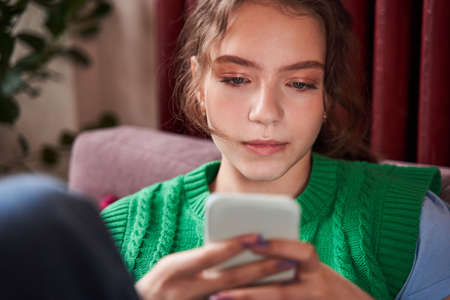 Caucasian girl sitting at the sofa and playing at the smartphone or reading messages at home