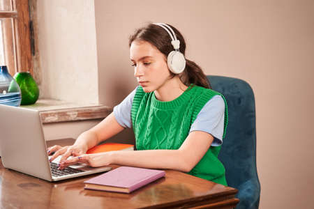 Female teen wearing headphones typing at the laptop computer while doing her homework