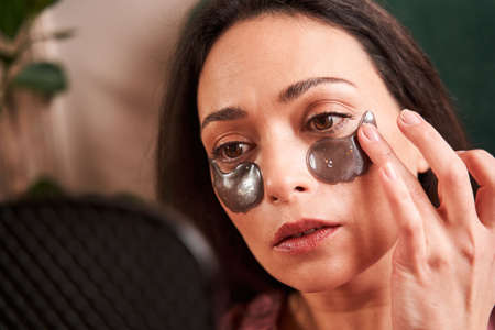 Woman with eye patches relaxing and looking at her reflection at the mirror