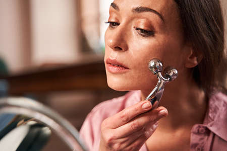 Woman is sitting in front of the mirror and looking at herself while using roller on cheeks