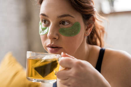 Caucasian woman with under eye patches drinking hot tea while relaxing at the sofaの写真素材