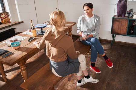 Female artist and her mature student drinking hot coffee and talking while sitting at the benchの写真素材