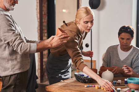 Female artist in apron choosing paints for plate and explaining how to drawing on itの写真素材