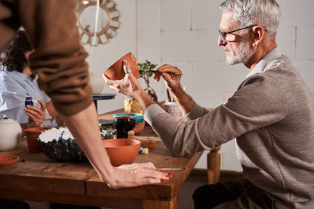Man wearing glasses drawing with paints at the clay pot while sitting at the master classの写真素材