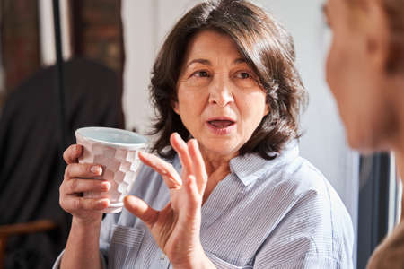 Mature woman holding beautiful cup of tea and talking with her young skilful teacherの写真素材