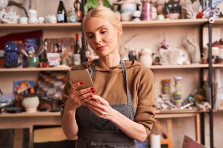 Woman wearing apron relaxing at her art studio and reading messages at the smartphoneの写真素材