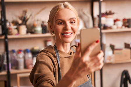 Cheerful female artist holding mobile phone and making self portrait on it at her art studioの写真素材