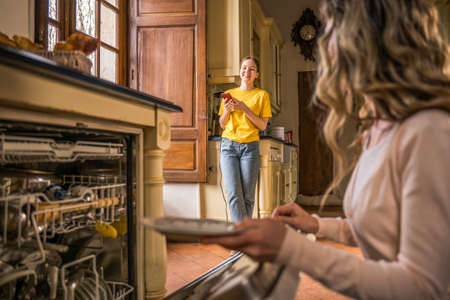 Mother putting dishes at the washing machine in kitchen while chatting with her teen child
