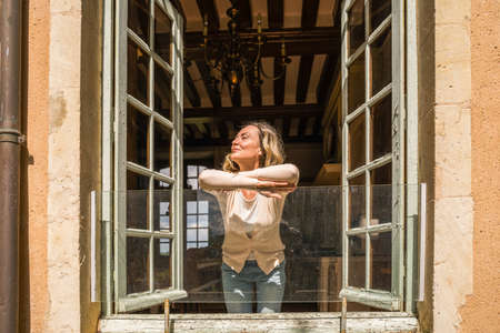 Adult woman enjoying sunshine at opened window during the summer day