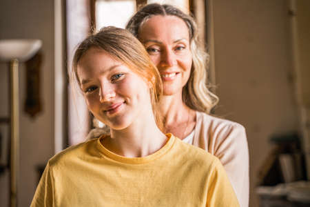 Mother looking at the camera while embracing her teen daughter at the kitchen