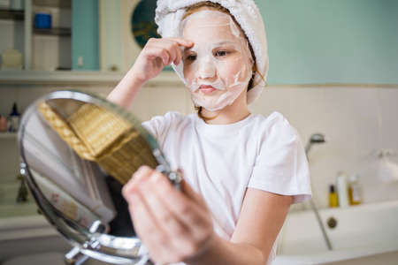 Female child holding hand mirror and looking at her reflection while applying face maskの写真素材