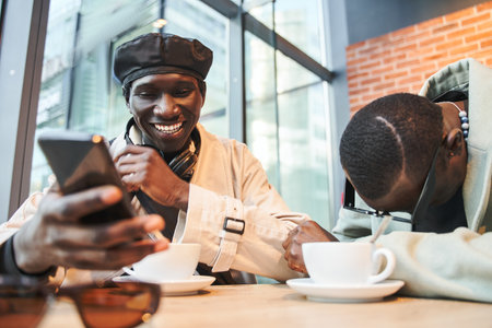 Smiling multiracial man talking to his friend in cozy cafe while looking at smartphoneの写真素材
