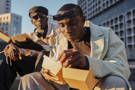 Trendy man eating his burger near male friend while sitting at the urban stairs