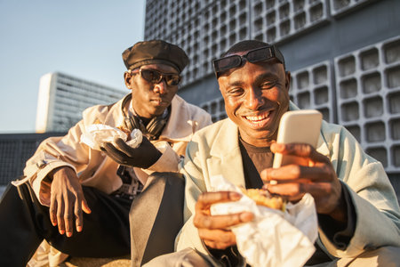 Cheerful multiracial hipster guys eating fast food and using smartphone while sitting on street stairsの写真素材