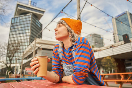 Smiling woman sitting with carton cup of coffee and looking awayの写真素材