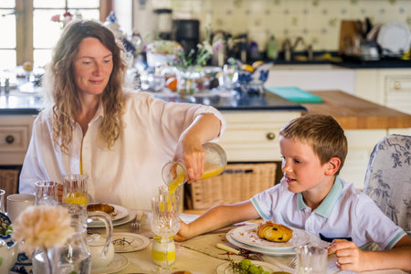 Young loving single mother pouring juice for her son while feeding him at kitchenの写真素材