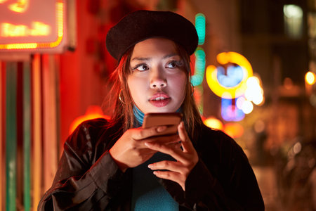 Young stylish female hipster standing near neon red light in night city and recording voice message