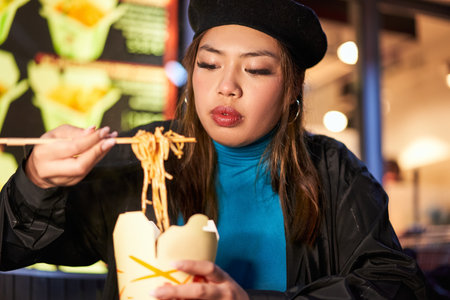 Asian woman eating noodles with sticks at the street cafe during the eveningの写真素材