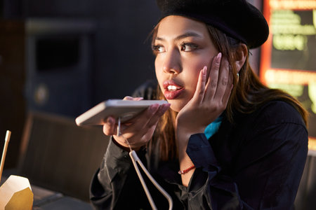 Stylish asian girl using phone while recording voice message at the illuminated cafeの写真素材