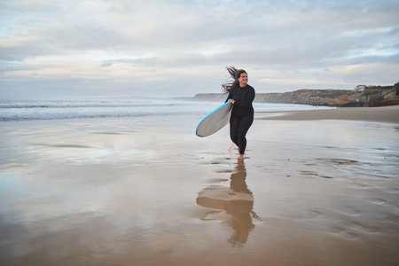 Cheerful surfer lady running at the sea shore and feeling happy during cloudy dayの写真素材