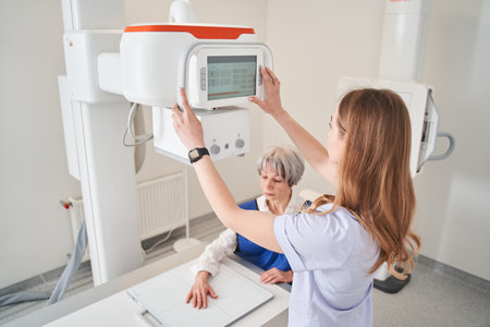 Woman doctor turning x ray apparatus while making arm examination to senior lady patientの写真素材