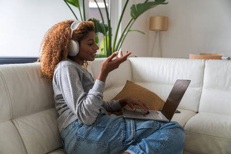 Positive woman wearing headphones working on laptop computer and talking at video callの写真素材