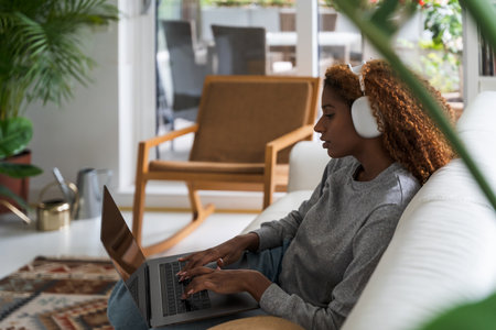 Curly young woman wearing headphones typing laptop computer keyboard at homeの写真素材