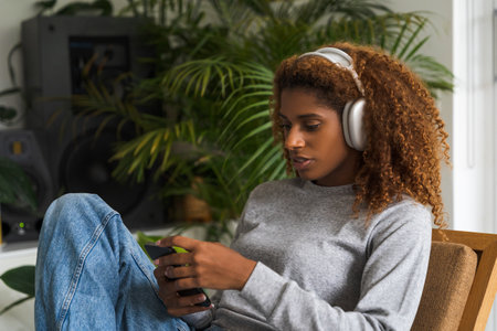 Positive curly woman wearing headphones listening music and looking at her smartphoneの写真素材