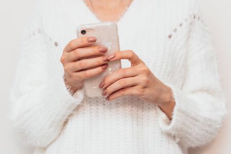 In the hands of a woman in a white sweater, a mobile phone in a very beautiful case. There is a neat beige manicure on the fingers.の写真素材