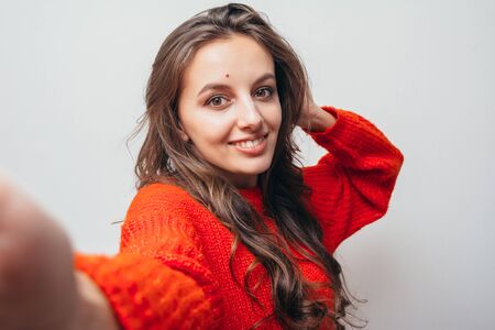 Beautiful girl with a mole takes a selfie in a red sweater against a white wall.
