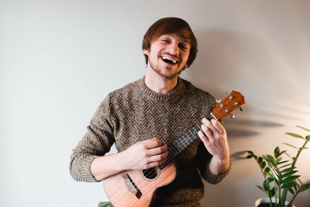 Man in a brown sweater smiles and plays on the ukulele while sitting at home.の写真素材