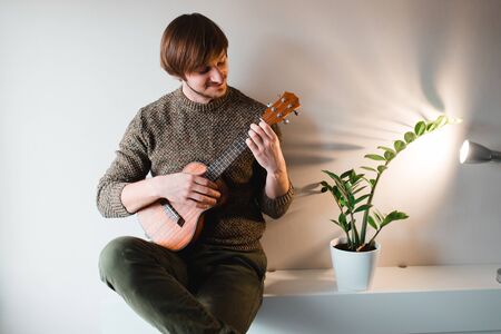 Man in a brown sweater smiles and plays on the ukulele while sitting at home.