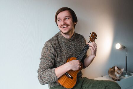 Man in a brown sweater smiles and plays on the ukulele while sitting at home.の写真素材