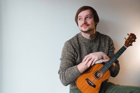 Man in a brown sweater smiles and plays on the ukulele while sitting at home.の写真素材