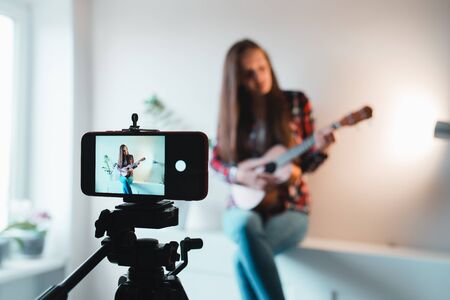 Girl in a shirt and jeans writes a vlog on the phone about playing ukulele.の写真素材