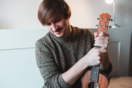 Man in a brown sweater smiles and plays on the ukulele while sitting at home.の写真素材