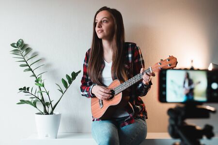 Girl in a shirt and jeans writes a vlog on the phone about playing ukulele.の写真素材