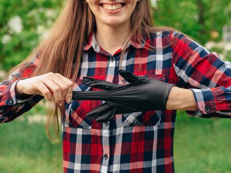 A young girl in a red checkered shirt takes off black latex black gloves on a background of green trees. End covid-2019.の写真素材