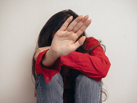 A girl in a red shirt and jeans sits sad against the wall. Domestic violence. She raised her hand asking for a stop.の写真素材