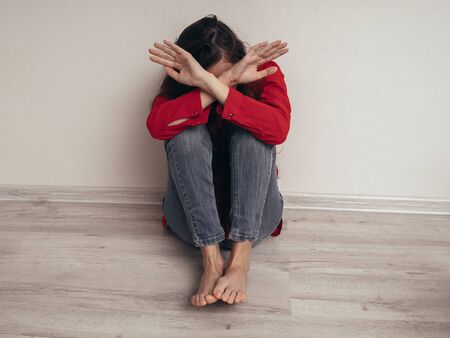 A girl in a red shirt and jeans sits sad against the wall. Domestic violence.の写真素材