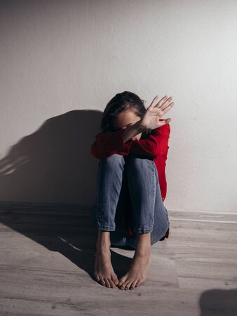 A girl in a red shirt and jeans sits sad against the wall. Domestic violence. She raised her hand asking for a stop.の写真素材