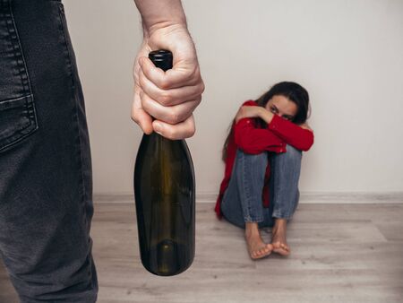 Male hand with a bottle of alcohol on the background of a frightened girl in a red shirt and jeans. Domestic violence. Alcoholism.の写真素材