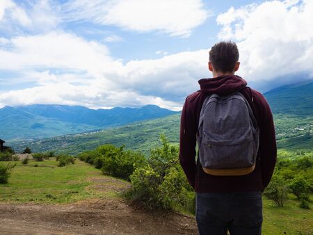 A young guy in a red hoodie and gray backpack travels in the mountains among green trees and clouds.