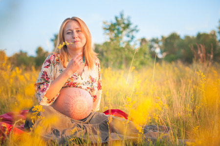 9 months pregnant woman with mehandi pattern ornament belly sitting in yellow grass and smiling on sunset lights. Waiting for baby. Pregnancy conceptの写真素材
