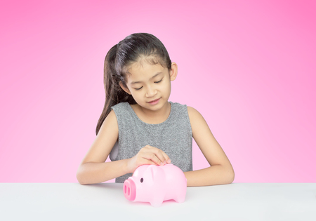Asian girl age around 8 years old putting coin into pink piggy bank on pink background with clipping pathの写真素材