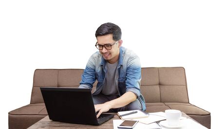 Asian guy surfing internet or using laptop sitting on sofa working as freelance at home   for graphic designの写真素材