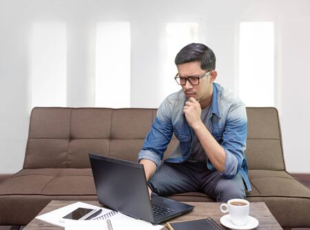 Asian guy surfing internet or using laptop sitting on sofa working as freelance at home   for graphic designの写真素材